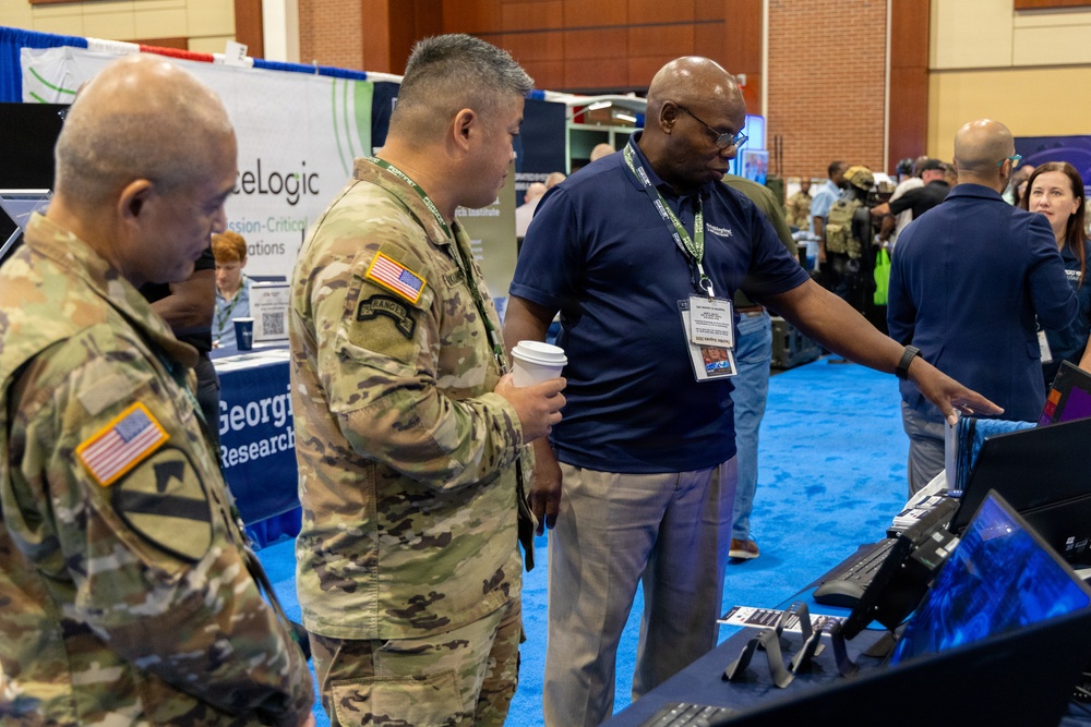 The image shows a military technology or defense expo/conference where uniformed U.S. Army soldiers and civilian personnel are gathered at vendor booths. A civilian representative wearing a lanyard badge is pointing at equipment on a display table, appearing to demonstrate products to the soldiers. The event takes place in a large convention hall with multiple vendor booths visible in the background.