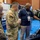The image shows a military technology or defense expo/conference where uniformed U.S. Army soldiers and civilian personnel are gathered at vendor booths. A civilian representative wearing a lanyard badge is pointing at equipment on a display table, appearing to demonstrate products to the soldiers. The event takes place in a large convention hall with multiple vendor booths visible in the background.