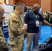 The image shows a military technology or defense expo/conference where uniformed U.S. Army soldiers and civilian personnel are gathered at vendor booths. A civilian representative wearing a lanyard badge is pointing at equipment on a display table, appearing to demonstrate products to the soldiers. The event takes place in a large convention hall with multiple vendor booths visible in the background.