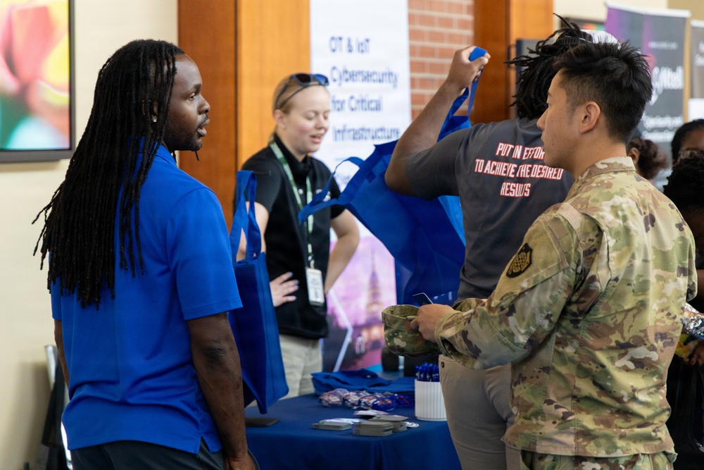 The image depicts what appears to be a career fair or expo event, where individuals are interacting at a booth focused on OT &amp; IoT Cybersecurity for Critical Infrastructure. A person in a blue polo shirt is speaking with someone in military camouflage uniform, while another attendee in a gray shirt holds up a blue tote bag. A booth representative wearing a lanyard badge is visible in the background.