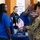 The image depicts what appears to be a career fair or expo event, where individuals are interacting at a booth focused on OT &amp; IoT Cybersecurity for Critical Infrastructure. A person in a blue polo shirt is speaking with someone in military camouflage uniform, while another attendee in a gray shirt holds up a blue tote bag. A booth representative wearing a lanyard badge is visible in the background.