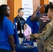The image depicts what appears to be a career fair or expo event, where individuals are interacting at a booth focused on OT &amp; IoT Cybersecurity for Critical Infrastructure. A person in a blue polo shirt is speaking with someone in military camouflage uniform, while another attendee in a gray shirt holds up a blue tote bag. A booth representative wearing a lanyard badge is visible in the background.