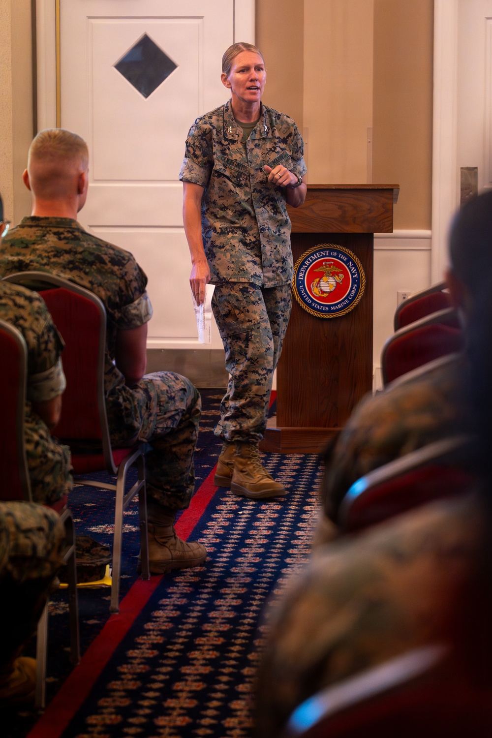 A female U.S. Marine Corps officer in MARPAT camouflage uniform is addressing an audience of seated Marines in a formal indoor setting. She is standing in front of a wooden podium bearing the United States Marine Corps seal (Department of the Navy). The setting appears to be a briefing, ceremony, or official presentation.