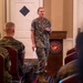 A female U.S. Marine Corps officer in MARPAT camouflage uniform is addressing an audience of seated Marines in a formal indoor setting. She is standing in front of a wooden podium bearing the United States Marine Corps seal (Department of the Navy). The setting appears to be a briefing, ceremony, or official presentation.