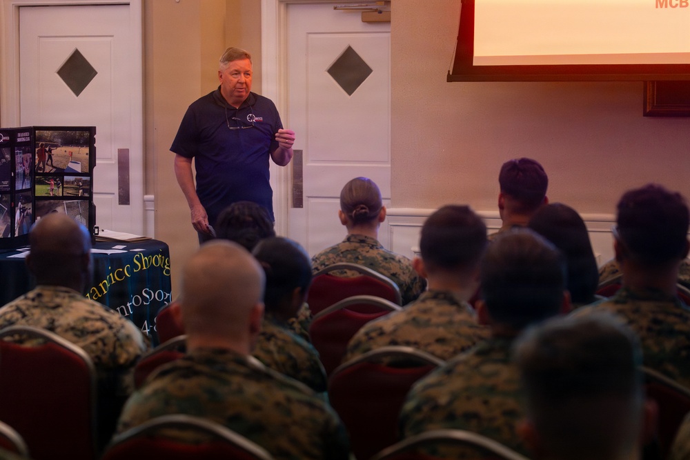 A civilian speaker addresses a large group of military personnel, likely U.S. Marines, in what appears to be a formal briefing or presentation room. The speaker, an older man in a dark polo shirt, stands at the front near a display board and a projection screen showing 'MCB'. The audience is seated in rows wearing camouflage uniforms.