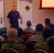 A civilian speaker addresses a large group of military personnel, likely U.S. Marines, in what appears to be a formal briefing or presentation room. The speaker, an older man in a dark polo shirt, stands at the front near a display board and a projection screen showing 'MCB'. The audience is seated in rows wearing camouflage uniforms.