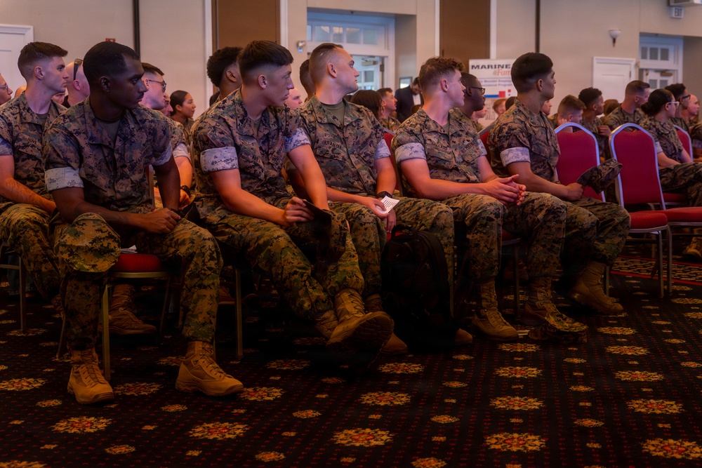 A large group of U.S. Marines in MARPAT camouflage uniforms are seated in rows of red chairs inside what appears to be a banquet hall or conference room. The Marines are attentively facing forward, suggesting they are attending a briefing, ceremony, or formal presentation. A partially visible sign in the background reads 'MARINE' and 'Fam', likely referencing a Marine Family event.