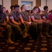 A large group of U.S. Marines in MARPAT camouflage uniforms are seated in rows of red chairs inside what appears to be a banquet hall or conference room. The Marines are attentively facing forward, suggesting they are attending a briefing, ceremony, or formal presentation. A partially visible sign in the background reads 'MARINE' and 'Fam', likely referencing a Marine Family event.