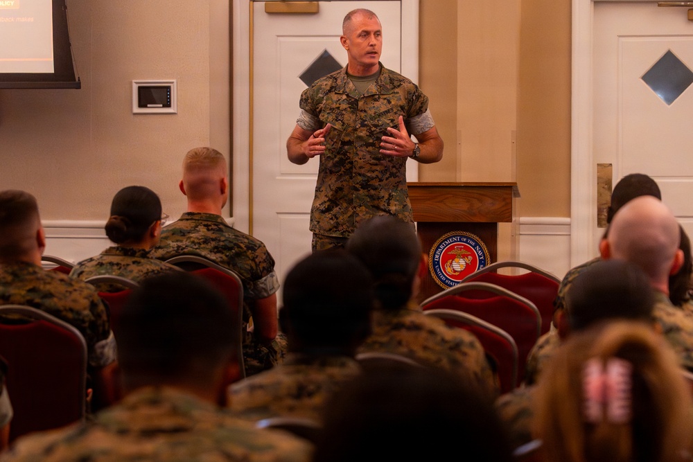 A U.S. Marine Corps officer in MARPAT camouflage uniform is addressing a room full of seated Marines at what appears to be a formal briefing or training event. A Department of the Navy seal is visible on the wooden podium beside him. The audience is seated in red chairs facing the speaker, with a presentation screen visible in the background.