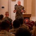 A U.S. Marine Corps officer in MARPAT camouflage uniform is addressing a room full of seated Marines at what appears to be a formal briefing or training event. A Department of the Navy seal is visible on the wooden podium beside him. The audience is seated in red chairs facing the speaker, with a presentation screen visible in the background.