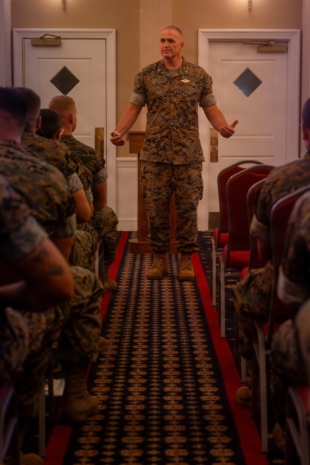 A senior military officer in MARPAT (Marine Pattern) camouflage uniform stands at the front of a room addressing a seated audience of fellow Marines. The speaker is positioned between two white doors in what appears to be a hotel ballroom or conference facility, gesturing with both hands while speaking. The audience is arranged in rows on either side of a central aisle.