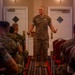 A senior military officer in MARPAT (Marine Pattern) camouflage uniform stands at the front of a room addressing a seated audience of fellow Marines. The speaker is positioned between two white doors in what appears to be a hotel ballroom or conference facility, gesturing with both hands while speaking. The audience is arranged in rows on either side of a central aisle.