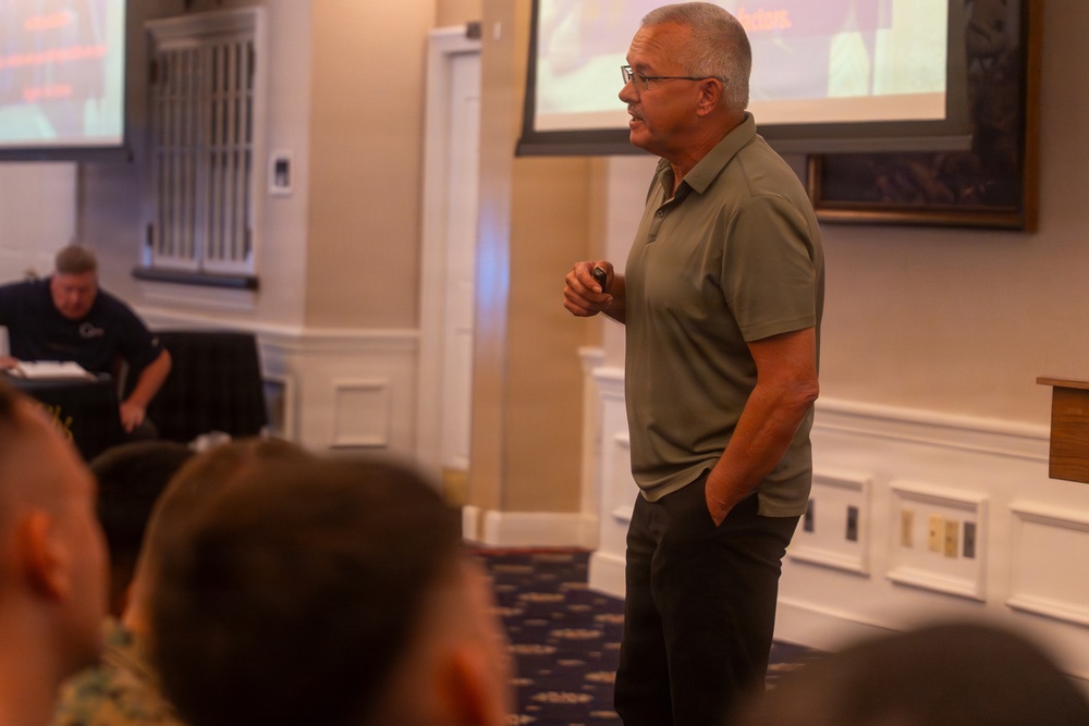 A middle-aged man wearing a green polo shirt and glasses is standing and presenting to an audience in a formal conference or banquet room. He is holding what appears to be a presentation clicker in one hand while addressing seated attendees. Two projection screens are visible in the background, displaying content related to the presentation.