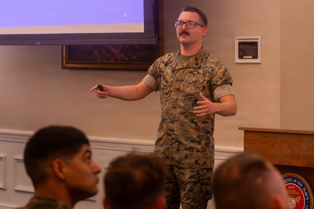 A U.S. Marine in MARPAT camouflage uniform is delivering a presentation to a seated audience in a formal briefing room. He is standing near a wooden podium bearing a Department of the Navy seal, gesturing with both hands while holding what appears to be a presentation clicker. A projection screen is visible in the background.