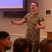 A U.S. Marine in MARPAT camouflage uniform is delivering a presentation to a seated audience in a formal briefing room. He is standing near a wooden podium bearing a Department of the Navy seal, gesturing with both hands while holding what appears to be a presentation clicker. A projection screen is visible in the background.