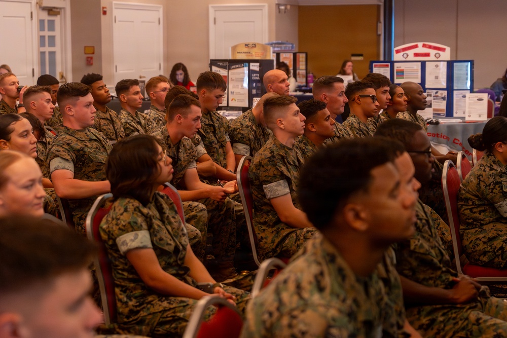 A large group of U.S. Marines in woodland MARPAT camouflage uniforms are seated in rows of red chairs, attending what appears to be a briefing or informational event. The background features multiple display boards and informational booths, suggesting an educational fair, resource event, or command briefing. The audience appears attentive and engaged.