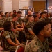 A large group of U.S. Marines in woodland MARPAT camouflage uniforms are seated in rows of red chairs, attending what appears to be a briefing or informational event. The background features multiple display boards and informational booths, suggesting an educational fair, resource event, or command briefing. The audience appears attentive and engaged.