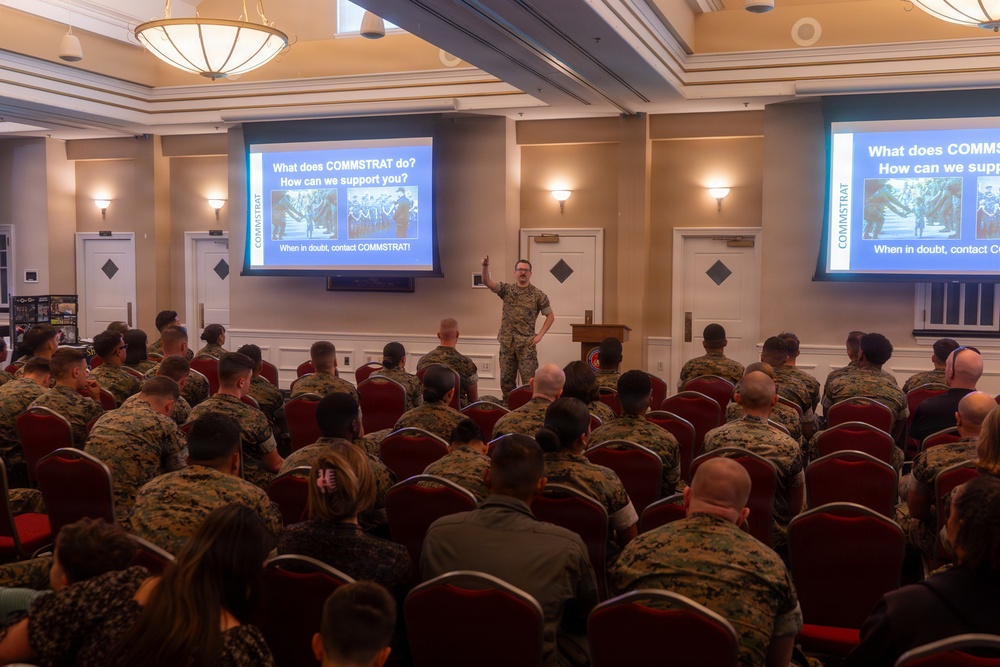 A military briefing or training session is taking place in a formal conference or banquet hall. A presenter in military camouflage uniform stands at the front, gesturing toward a projected slide that reads 'What does COMMSTRAT do? How can we support you?' The audience consists predominantly of personnel wearing U.S. Marine Corps MARPAT camouflage uniforms, seated in rows of red chairs.