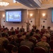 A military briefing or training session is taking place in a formal conference or banquet hall. A presenter in military camouflage uniform stands at the front, gesturing toward a projected slide that reads 'What does COMMSTRAT do? How can we support you?' The audience consists predominantly of personnel wearing U.S. Marine Corps MARPAT camouflage uniforms, seated in rows of red chairs.