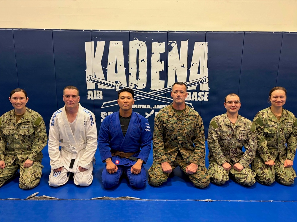 Six military personnel are posing together on a blue mat at Kadena Air Base in Okinawa, Japan. Two individuals in the center are wearing Brazilian Jiu-Jitsu gis (one white, one blue), while the others are in military camouflage uniforms. The setting appears to be a martial arts training facility or dojo on the base.
