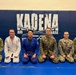 Six military personnel are posing together on a blue mat at Kadena Air Base in Okinawa, Japan. Two individuals in the center are wearing Brazilian Jiu-Jitsu gis (one white, one blue), while the others are in military camouflage uniforms. The setting appears to be a martial arts training facility or dojo on the base.