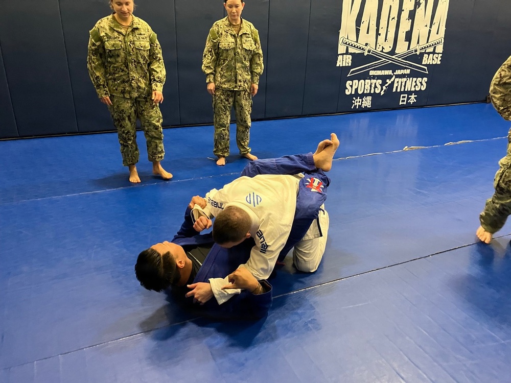 A Brazilian Jiu-Jitsu training session is taking place at Kadena Air Base Sports &amp; Fitness facility in Okinawa, Japan. Two practitioners in gi uniforms are grappling on a blue mat while military personnel in camouflage uniforms observe. The setting appears to be an official military base martial arts training event.