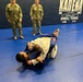 A Brazilian Jiu-Jitsu training session is taking place at Kadena Air Base Sports &amp; Fitness facility in Okinawa, Japan. Two practitioners in gi uniforms are grappling on a blue mat while military personnel in camouflage uniforms observe. The setting appears to be an official military base martial arts training event.