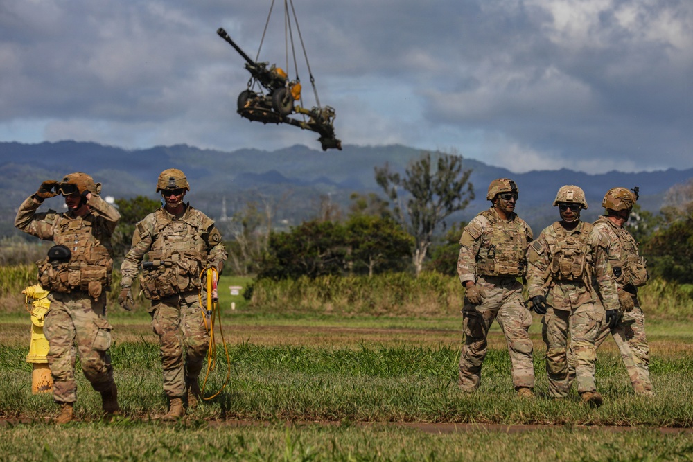 A group of five military personnel in U.S. Army OCP (Operational Camouflage Pattern) uniforms are standing on a grassy field during what appears to be a sling-load training exercise. An artillery piece, likely an M119 howitzer, is suspended in the air by a sling-load cable attached to an unseen helicopter above. The setting appears to be a tropical or subtropical location, consistent with Hawaii, based on the mountainous green terrain in the background.