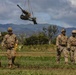 A group of five military personnel in U.S. Army OCP (Operational Camouflage Pattern) uniforms are standing on a grassy field during what appears to be a sling-load training exercise. An artillery piece, likely an M119 howitzer, is suspended in the air by a sling-load cable attached to an unseen helicopter above. The setting appears to be a tropical or subtropical location, consistent with Hawaii, based on the mountainous green terrain in the background.