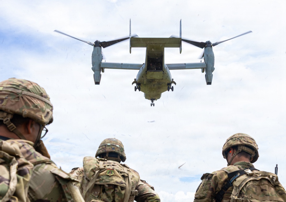 Three soldiers in full combat gear and helmets observe a Bell Boeing V-22 Osprey tiltrotor aircraft hovering directly overhead at low altitude. Debris and vegetation are visibly being blown around by the rotor wash. The scene appears to be a military training exercise or tactical insertion/extraction operation in an open field.