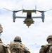 Three soldiers in full combat gear and helmets observe a Bell Boeing V-22 Osprey tiltrotor aircraft hovering directly overhead at low altitude. Debris and vegetation are visibly being blown around by the rotor wash. The scene appears to be a military training exercise or tactical insertion/extraction operation in an open field.