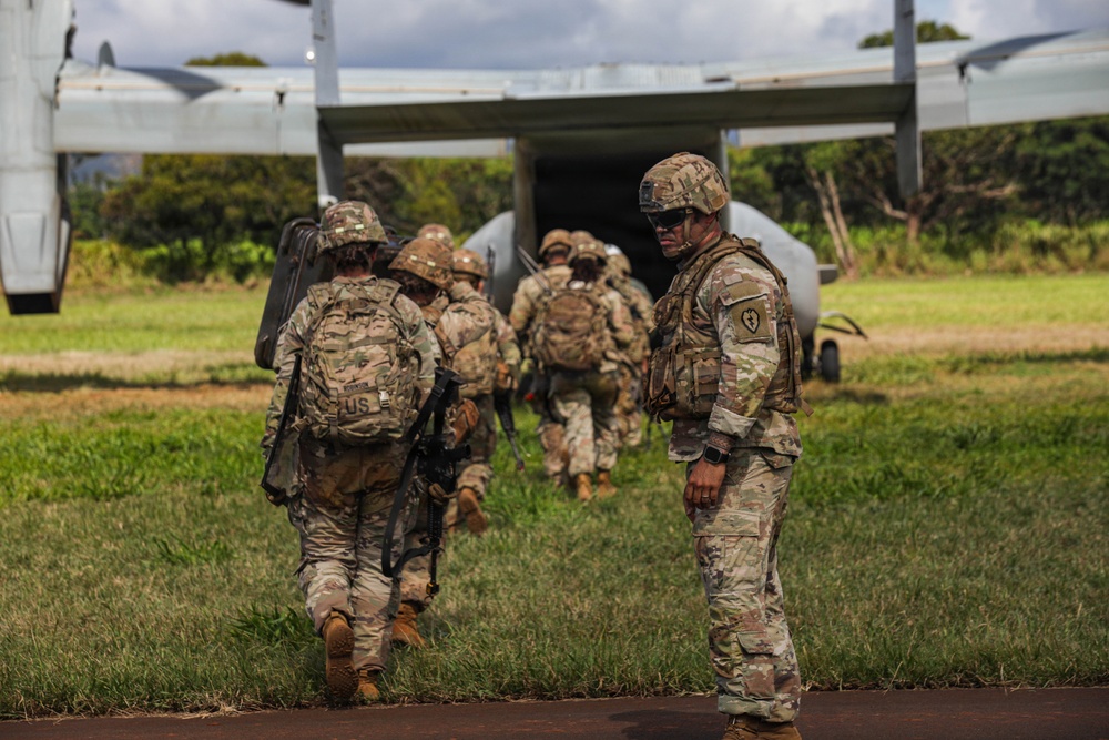 A group of U.S. Army soldiers in MultiCam/OCP camouflage uniforms are boarding a military tiltrotor aircraft, likely a V-22 Osprey, via its rear ramp. One soldier stands to the right facing the camera, wearing a unit patch on his left shoulder, while the others proceed toward the aircraft. The setting appears to be a grassy field or landing zone in a tropical or subtropical environment.