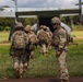 A group of U.S. Army soldiers in MultiCam/OCP camouflage uniforms are boarding a military tiltrotor aircraft, likely a V-22 Osprey, via its rear ramp. One soldier stands to the right facing the camera, wearing a unit patch on his left shoulder, while the others proceed toward the aircraft. The setting appears to be a grassy field or landing zone in a tropical or subtropical environment.