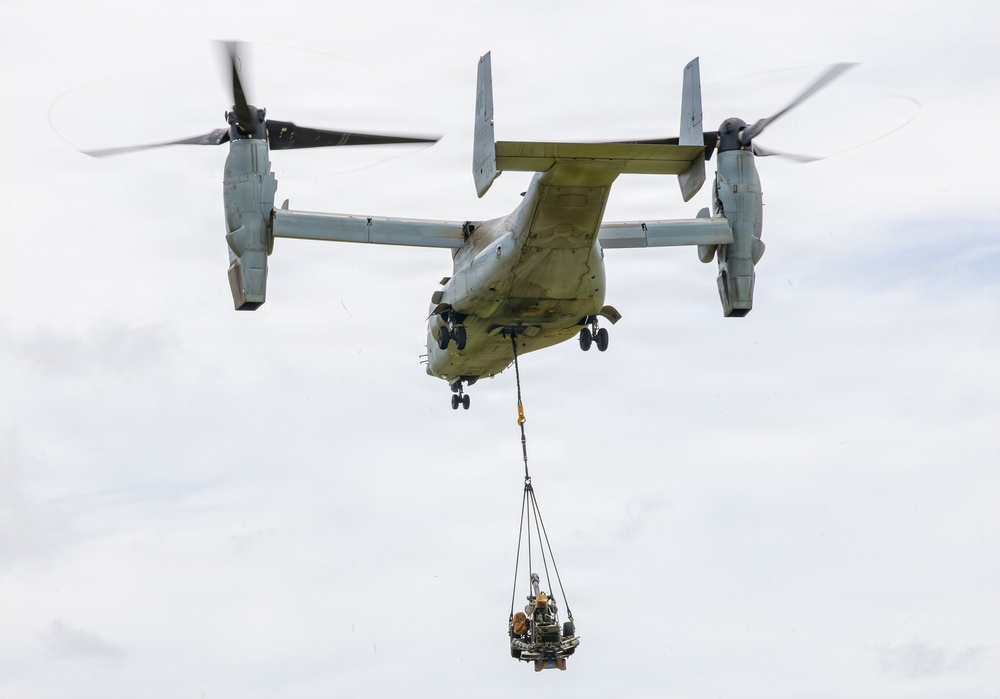 A tiltrotor aircraft, identifiable as a Bell Boeing V-22 Osprey, is captured in flight with its rotors in helicopter mode. The aircraft is performing an external sling load operation, carrying a piece of equipment or machinery suspended beneath its fuselage via a cargo net and rigging system. The overcast sky provides a neutral background, and the image is sharp and well-composed.