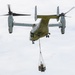 A tiltrotor aircraft, identifiable as a Bell Boeing V-22 Osprey, is captured in flight with its rotors in helicopter mode. The aircraft is performing an external sling load operation, carrying a piece of equipment or machinery suspended beneath its fuselage via a cargo net and rigging system. The overcast sky provides a neutral background, and the image is sharp and well-composed.