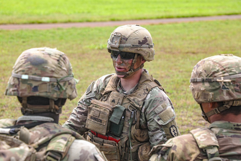 A U.S. Army Sergeant named Harrison, wearing OCP (Operational Camouflage Pattern) uniform with tactical gear and face camouflage paint, appears to be conducting a briefing or debriefing with fellow soldiers in an outdoor field setting. The central soldier displays Sergeant rank insignia on the chest rig and a unit patch on the left shoulder. The scene suggests a training exercise or field operation.