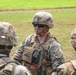 A U.S. Army Sergeant named Harrison, wearing OCP (Operational Camouflage Pattern) uniform with tactical gear and face camouflage paint, appears to be conducting a briefing or debriefing with fellow soldiers in an outdoor field setting. The central soldier displays Sergeant rank insignia on the chest rig and a unit patch on the left shoulder. The scene suggests a training exercise or field operation.