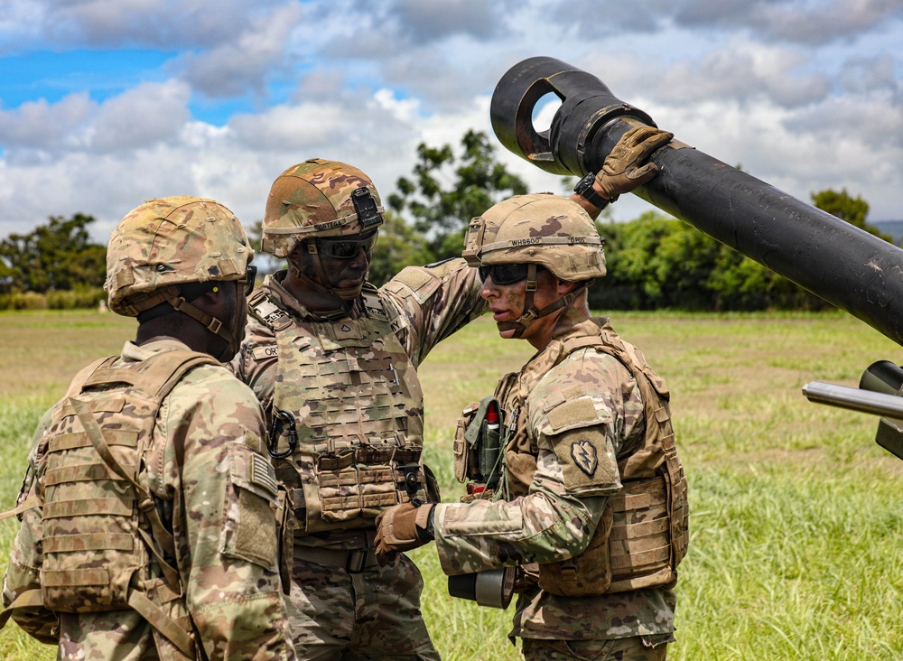Three military personnel in full combat gear are gathered around what appears to be a mortar or artillery piece in an open field. They appear to be engaged in a briefing, inspection, or training exercise, with one soldier placing a hand on another's helmet. The setting is a grassy outdoor area with trees and a partly cloudy sky in the background.