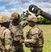 Three military personnel in full combat gear are gathered around what appears to be a mortar or artillery piece in an open field. They appear to be engaged in a briefing, inspection, or training exercise, with one soldier placing a hand on another's helmet. The setting is a grassy outdoor area with trees and a partly cloudy sky in the background.