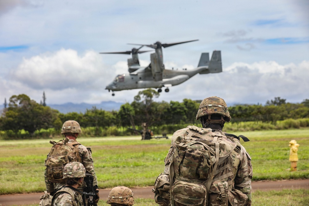 Military personnel in full combat gear observe a V-22 Osprey tiltrotor aircraft during what appears to be a training exercise or operational insertion. The soldiers are equipped with tactical backpacks and helmets, positioned on a grassy field with tropical vegetation in the background. The setting suggests a Pacific region location based on the lush green landscape and mountainous terrain.