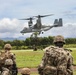 Military personnel in full combat gear observe a V-22 Osprey tiltrotor aircraft during what appears to be a training exercise or operational insertion. The soldiers are equipped with tactical backpacks and helmets, positioned on a grassy field with tropical vegetation in the background. The setting suggests a Pacific region location based on the lush green landscape and mountainous terrain.