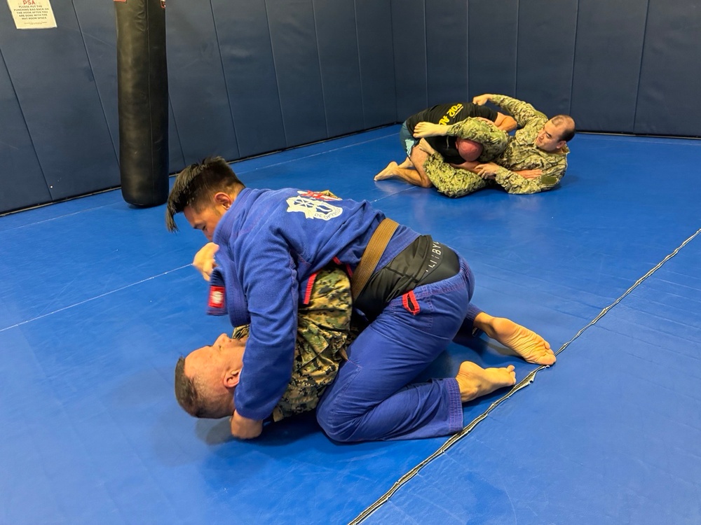 The image shows a Brazilian Jiu-Jitsu (BJJ) training session taking place on a blue mat in what appears to be a martial arts gym or military training facility. In the foreground, a practitioner in a blue gi with a brown belt is grappling with an opponent wearing military camouflage (MARPAT) pants. In the background, two additional individuals in military camouflage uniforms are also engaged in ground grappling.