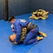 The image shows a Brazilian Jiu-Jitsu (BJJ) training session taking place on a blue mat in what appears to be a martial arts gym or military training facility. In the foreground, a practitioner in a blue gi with a brown belt is grappling with an opponent wearing military camouflage (MARPAT) pants. In the background, two additional individuals in military camouflage uniforms are also engaged in ground grappling.