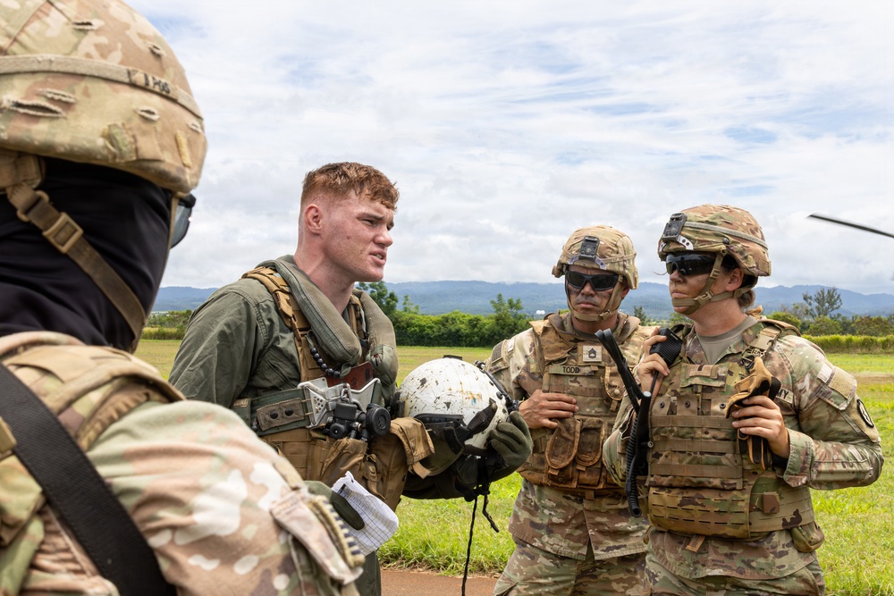 Four military personnel in OCP (Operational Camouflage Pattern) uniforms are engaged in a briefing or debrief on an open field with mountains visible in the background. One individual, wearing a flight suit and holding a white aviation helmet, appears to be a pilot or aircrew member speaking with ground soldiers. The setting suggests a landing zone or airfield area during a training exercise or operational mission.