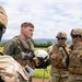 Four military personnel in OCP (Operational Camouflage Pattern) uniforms are engaged in a briefing or debrief on an open field with mountains visible in the background. One individual, wearing a flight suit and holding a white aviation helmet, appears to be a pilot or aircrew member speaking with ground soldiers. The setting suggests a landing zone or airfield area during a training exercise or operational mission.