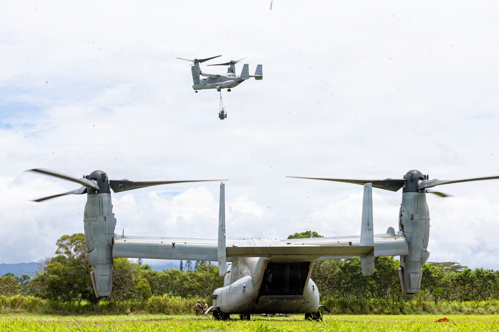 Two V-22 Osprey tiltrotor aircraft are visible in a tropical field setting. One Osprey is grounded on a grass landing zone with its rear ramp open and rotors spinning, while a second Osprey hovers overhead conducting an external sling load operation with a suspended cargo load. The lush green vegetation and mountainous background suggest a tropical or Pacific island environment.