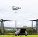 Two V-22 Osprey tiltrotor aircraft are visible in a tropical field setting. One Osprey is grounded on a grass landing zone with its rear ramp open and rotors spinning, while a second Osprey hovers overhead conducting an external sling load operation with a suspended cargo load. The lush green vegetation and mountainous background suggest a tropical or Pacific island environment.