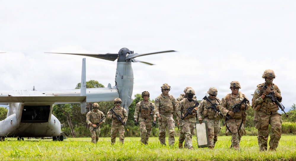 A group of approximately eight military personnel in full combat gear are disembarking from a V-22 Osprey tiltrotor aircraft on a grassy field. The soldiers are armed with assault rifles and equipped with tactical vests, helmets, and eye protection. The setting appears to be a tropical or subtropical environment based on the lush green vegetation visible in the background.