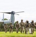 A group of approximately eight military personnel in full combat gear are disembarking from a V-22 Osprey tiltrotor aircraft on a grassy field. The soldiers are armed with assault rifles and equipped with tactical vests, helmets, and eye protection. The setting appears to be a tropical or subtropical environment based on the lush green vegetation visible in the background.