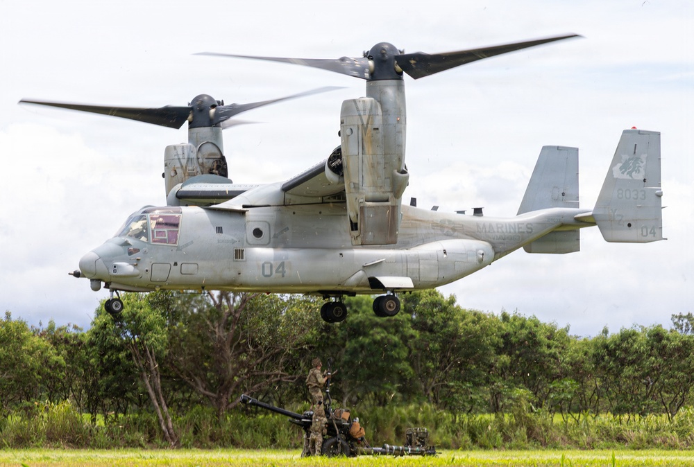 A U.S. Marines MV-22 Osprey tiltrotor aircraft (bureau number 8033, side number 04) hovers at low altitude over a grassy field. A soldier in camouflage uniform stands beneath or near the aircraft next to what appears to be an artillery piece or heavy weapon system. The scene suggests a military training exercise or sling-load/fire support coordination operation in a tropical or subtropical environment.